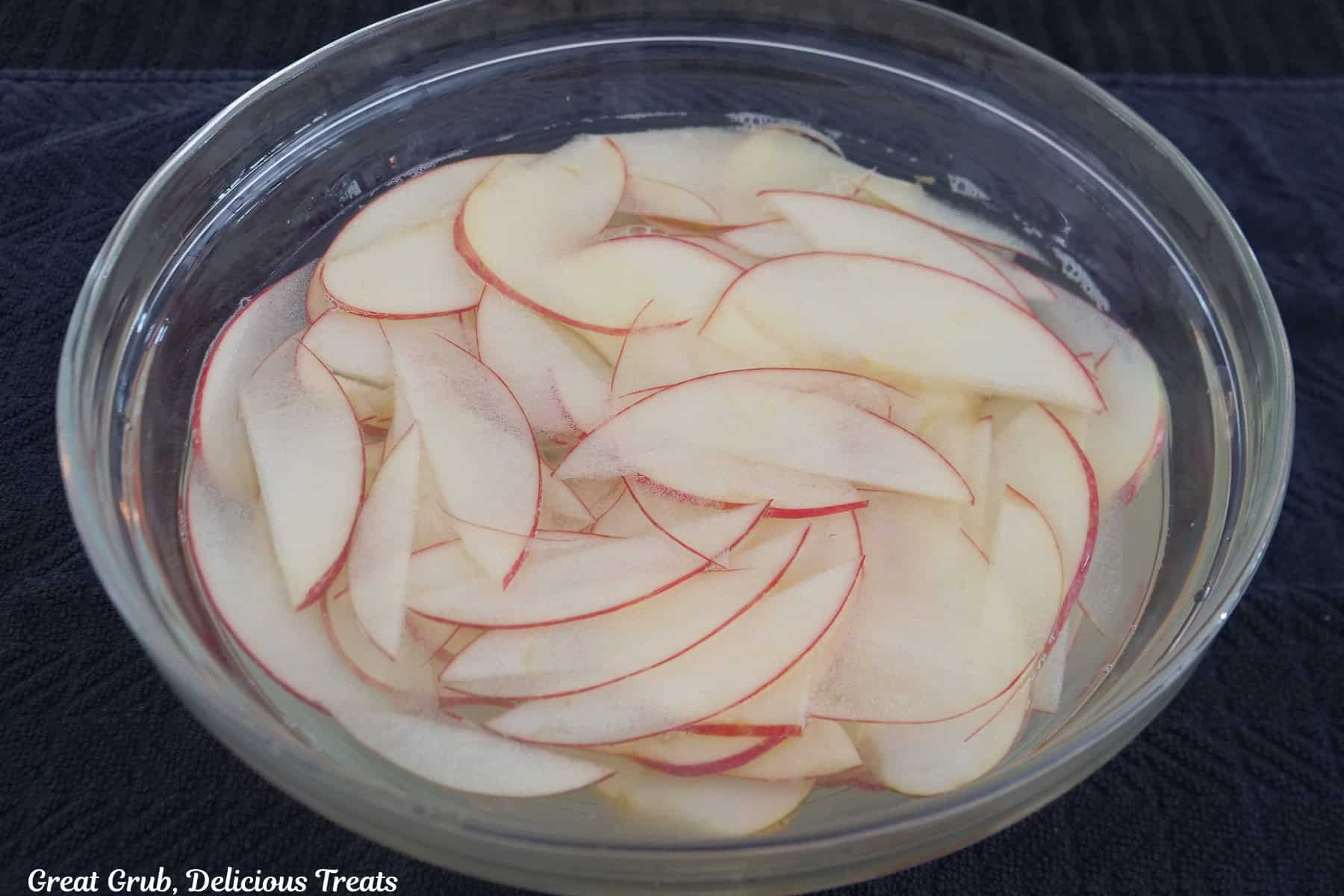 A glass bowl filled water and very thin apple slices soaking in it.
