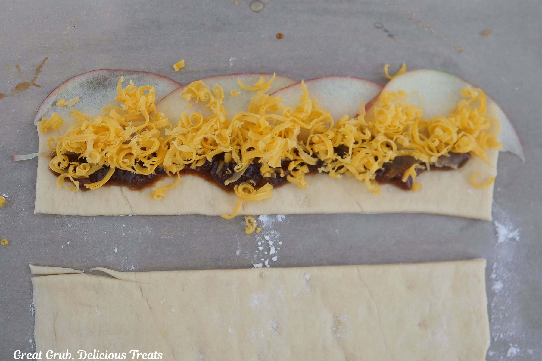 A pastry strip with bacon jam, thinly sliced apples, and shredded cheddar cheese on it before being rolled up into a pastry rose.