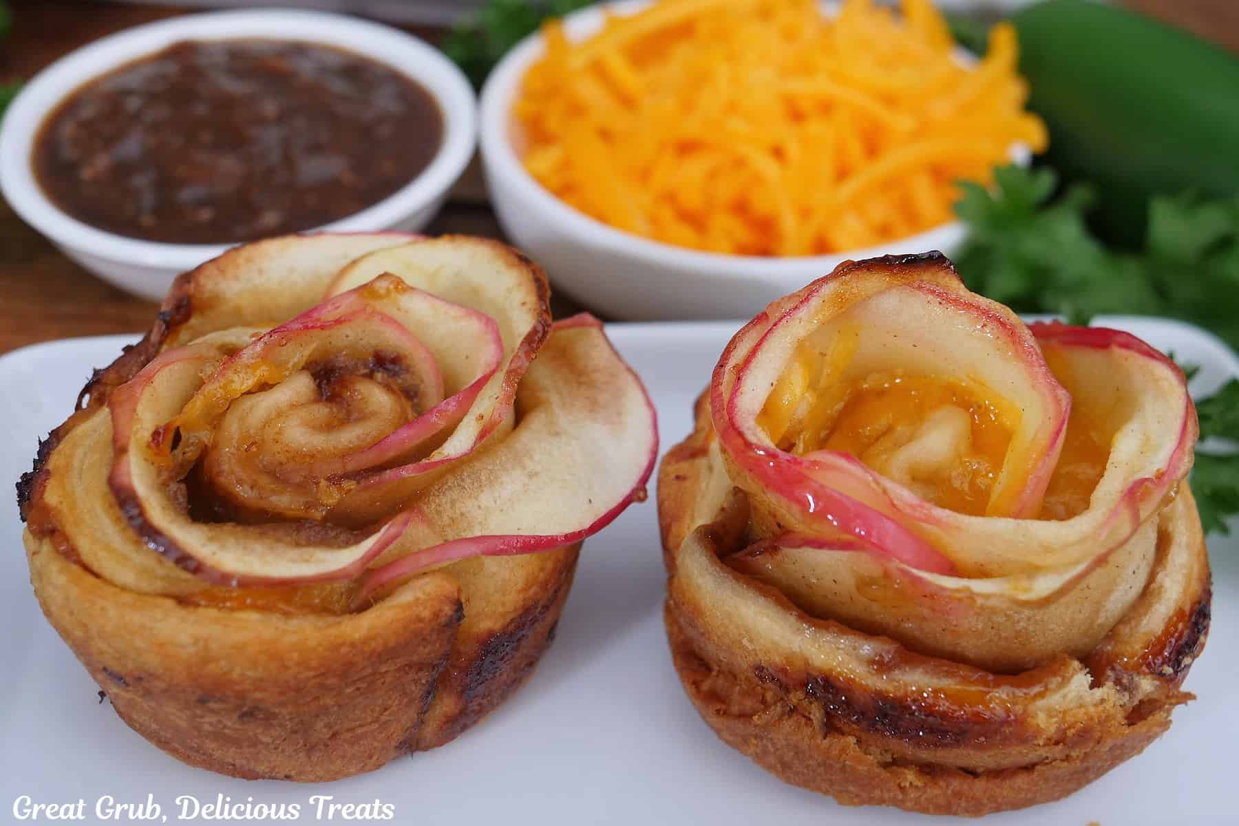 An oblong white plate with two cheesy apple bacon jam roses on it with two small white bowls in the background with cheese and bacon jam in them.