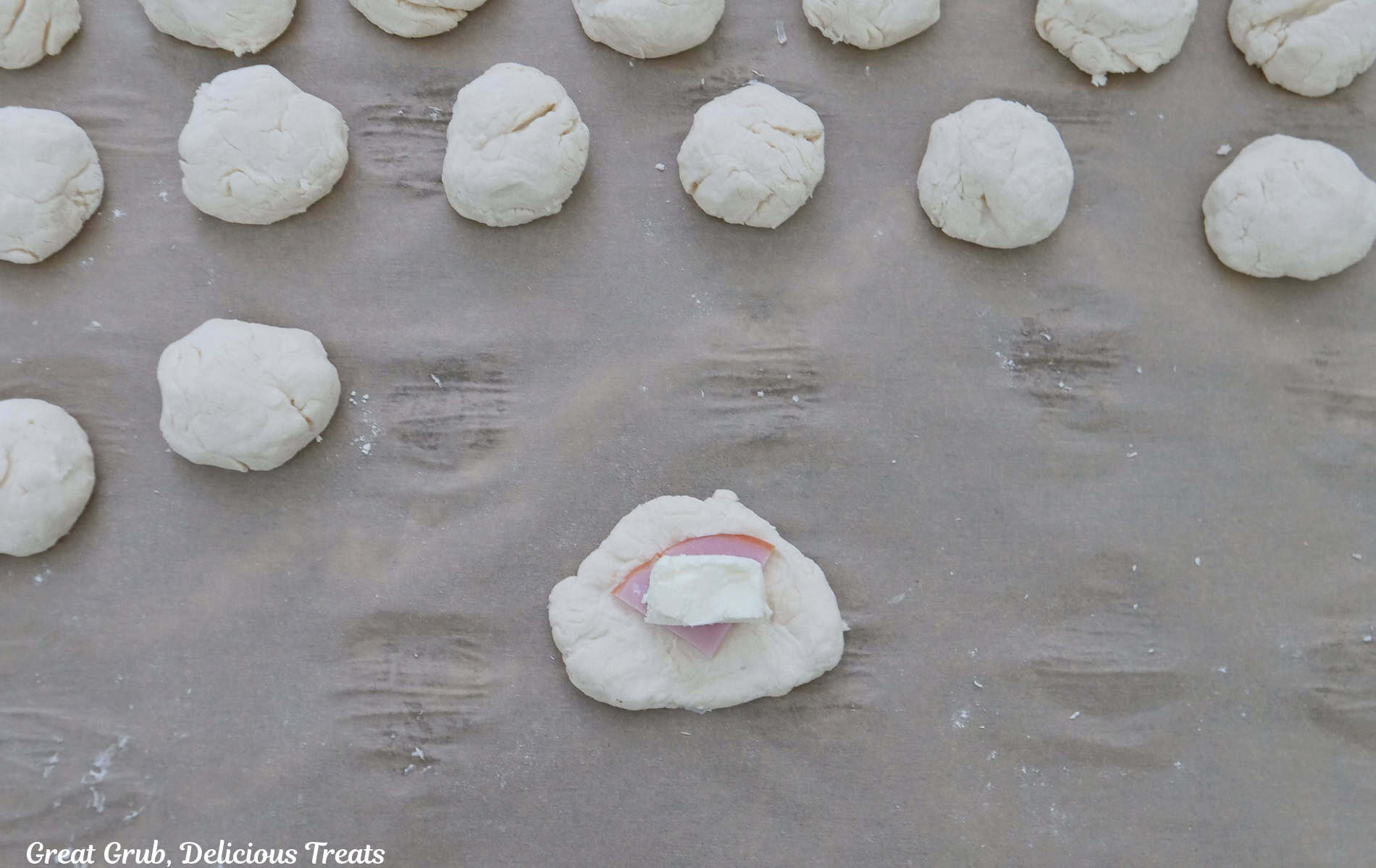 Parchment paper with dough balls on it with one flattened out with a chunk of cream cheese and a piece of Canadian bacon on it.
