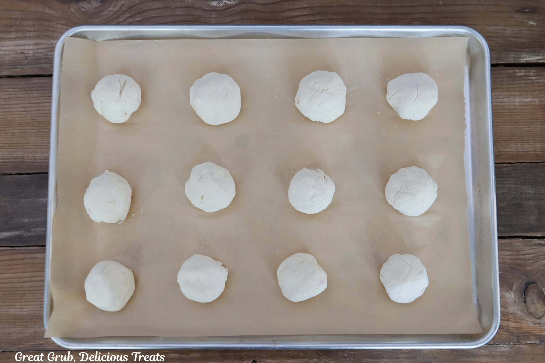 A wood surface with a baking sheet lined with parchment paper with sixteen dough balls on it.