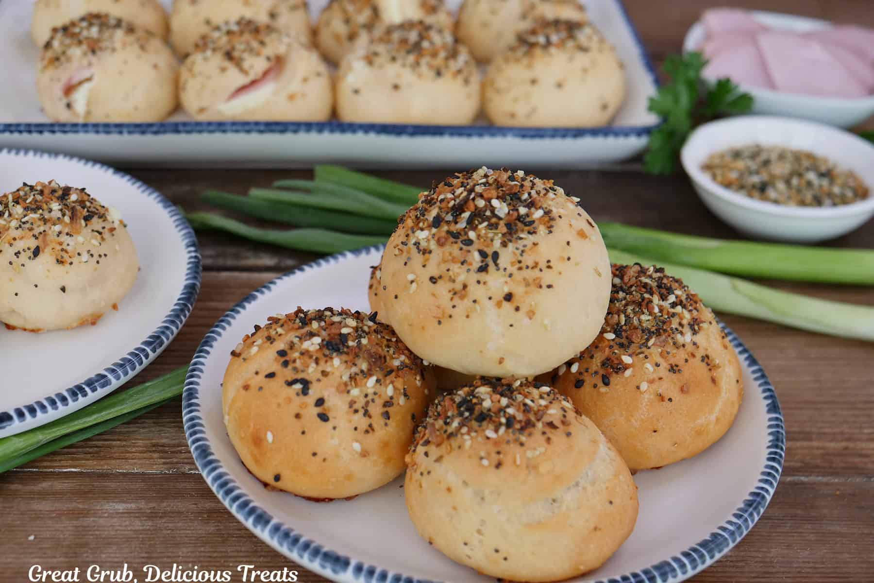 A wood surface with three white plates with blue trim with everything bagel bombs on them with Canadian bacon, green onions, and seasoning in the background.
