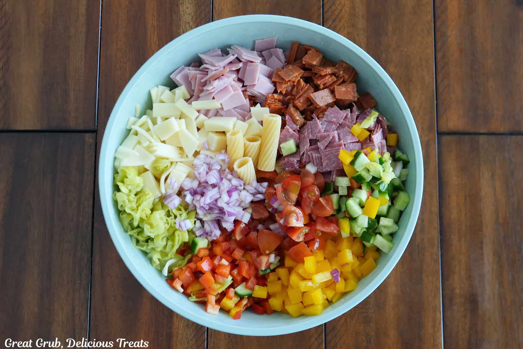 A wood surface with a light blue bowl filled with all the chopped ingredients for the grinder pasta salad in it.