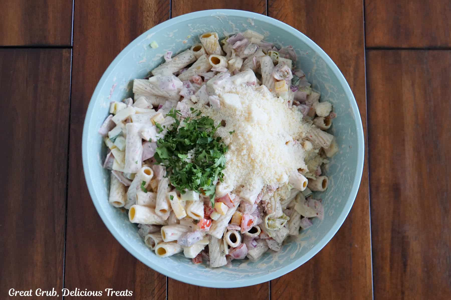 A light blue mixing bowl on a wood surface with the pasta ingredients mixed together and grated Parmesan and parsley on top before being mixed together.