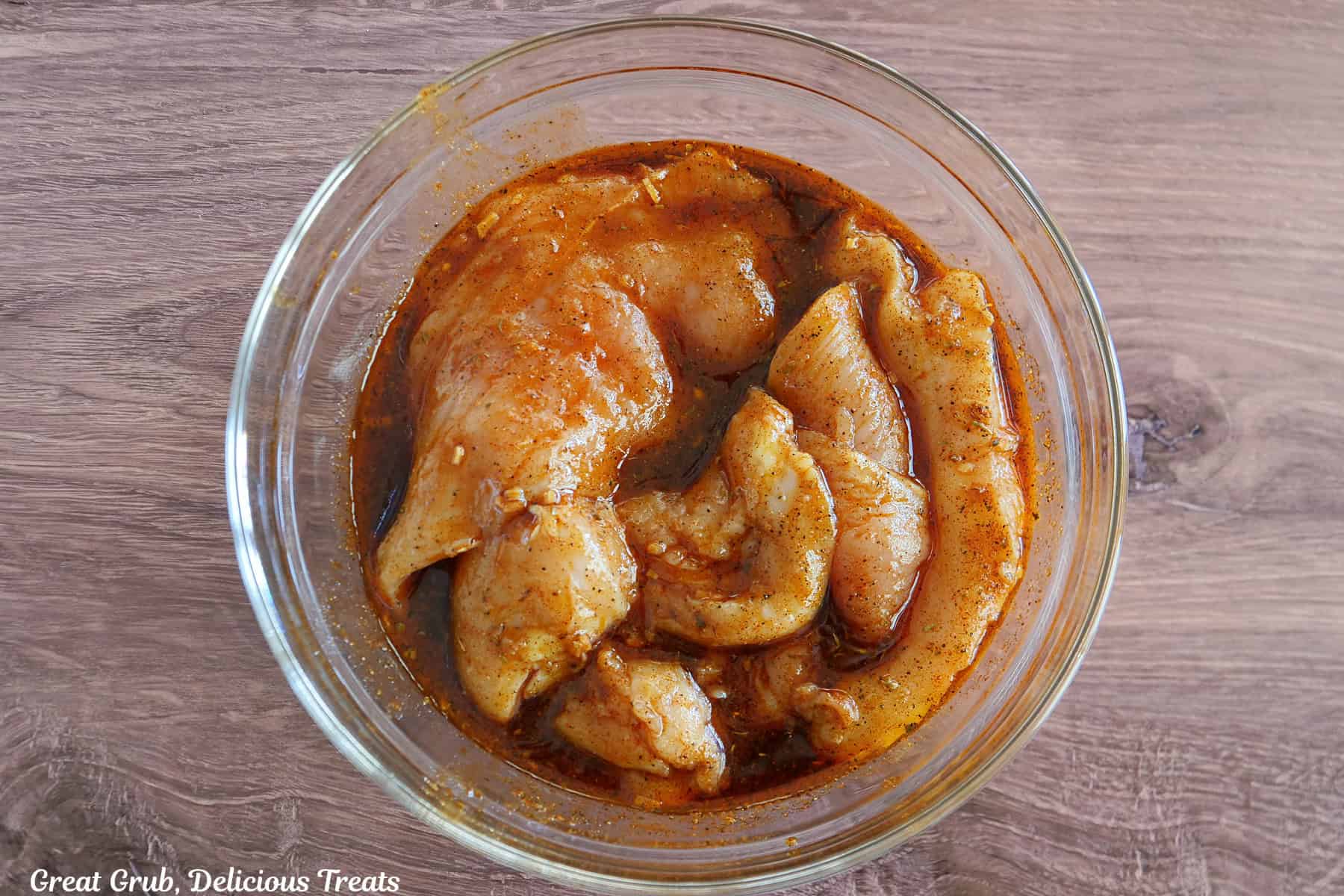 A wood surface with a glass bowl filled with marinating chicken breasts.