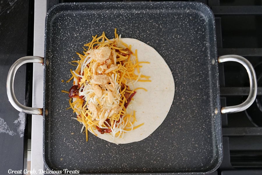 A square frying pan with a flour tortilla that has to types of cheese, bbq pulled pork and crispy fried onions on one half of the tortilla, being fried.