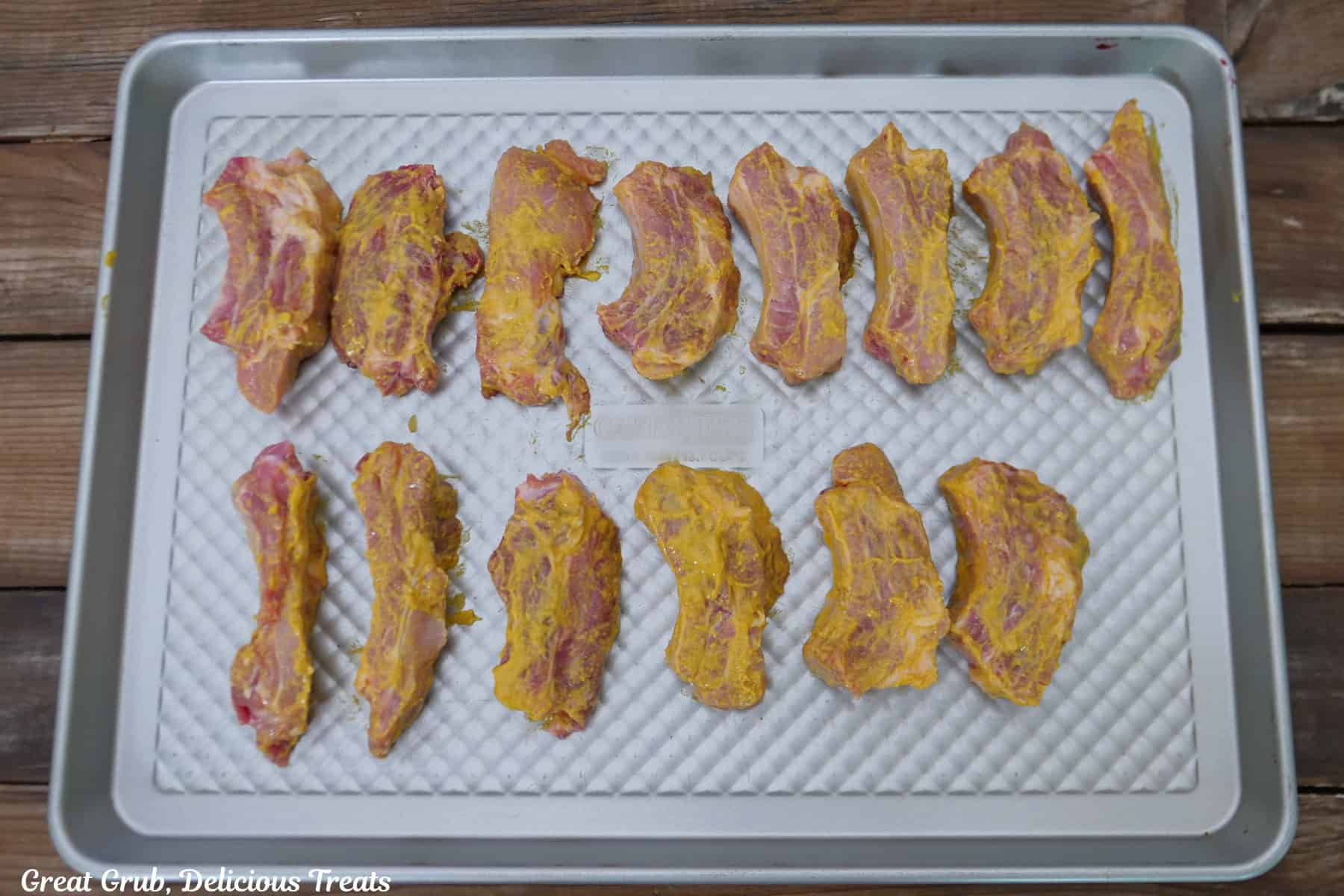 A sliver baking sheet with fourteen pork ribs that are covered with mustard.