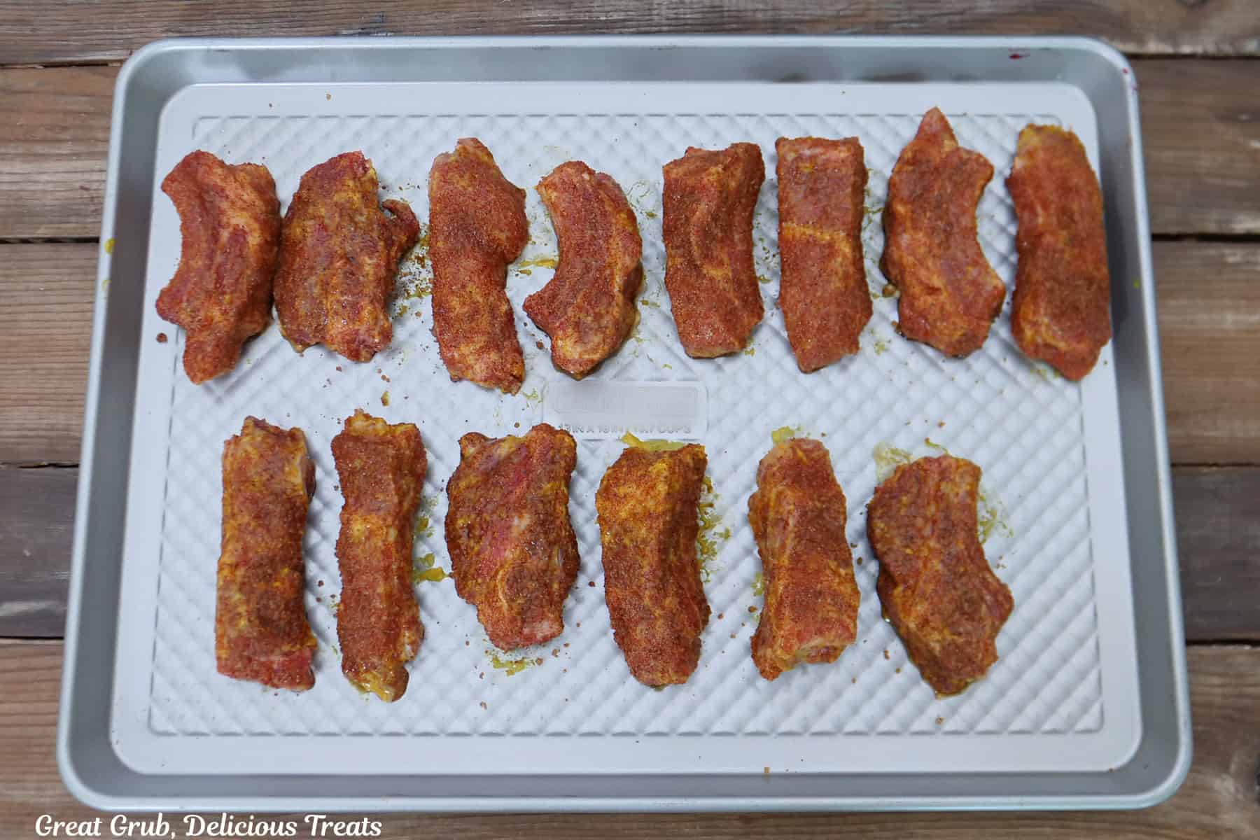 A sliver baking sheet with fourteen pork ribs that are covered with mustard and a zesty blend of seasonings.