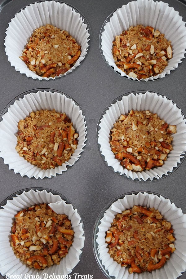 A jumbo muffin tin with white paper liners in it with pretzel crust ingredients pressed into the bottom of each liner.