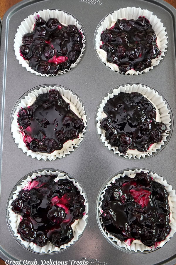 A jumbo muffin tin with mini blueberry pretzel desserts being assembled, with the sweet cream cheese mixture on top of the pretzel crusts, and blueberry topping on top of the cream cheese mixture.