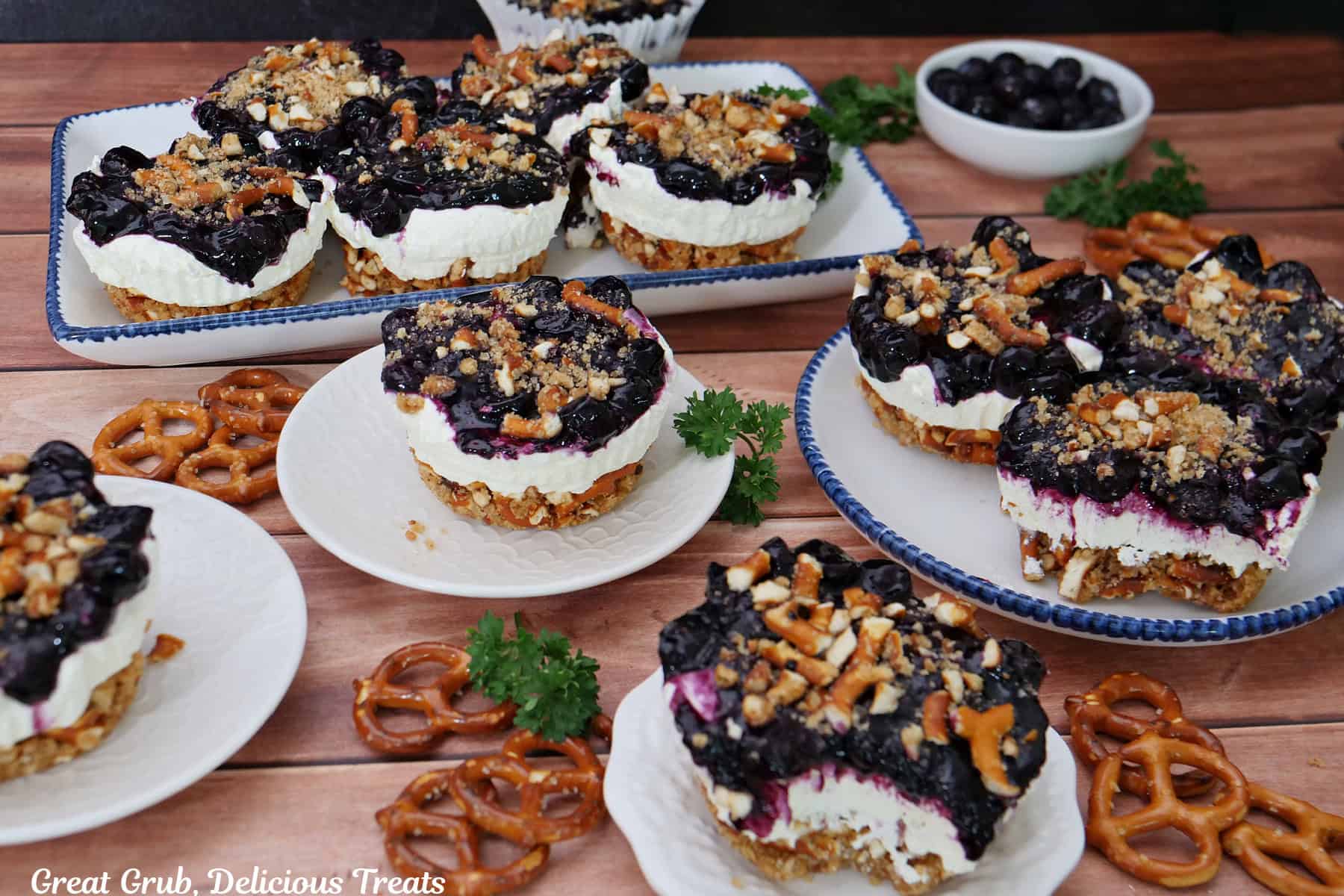 A wood surface with 12 mini blueberry pretzel desserts placed on white plate randomly placed around on the wood surface.