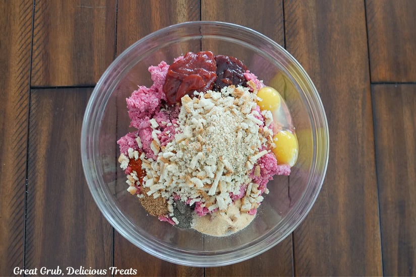 A wood surface with all the ingredients to mix together to make meatloaf in a glass bowl before being mixed together.