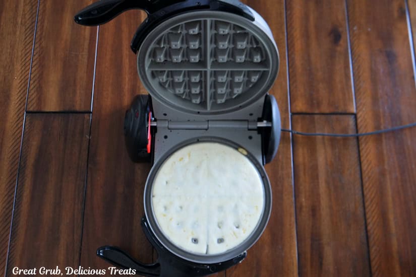 A wood surface with a waffle iron on it and waffle batter in the waffle maker.