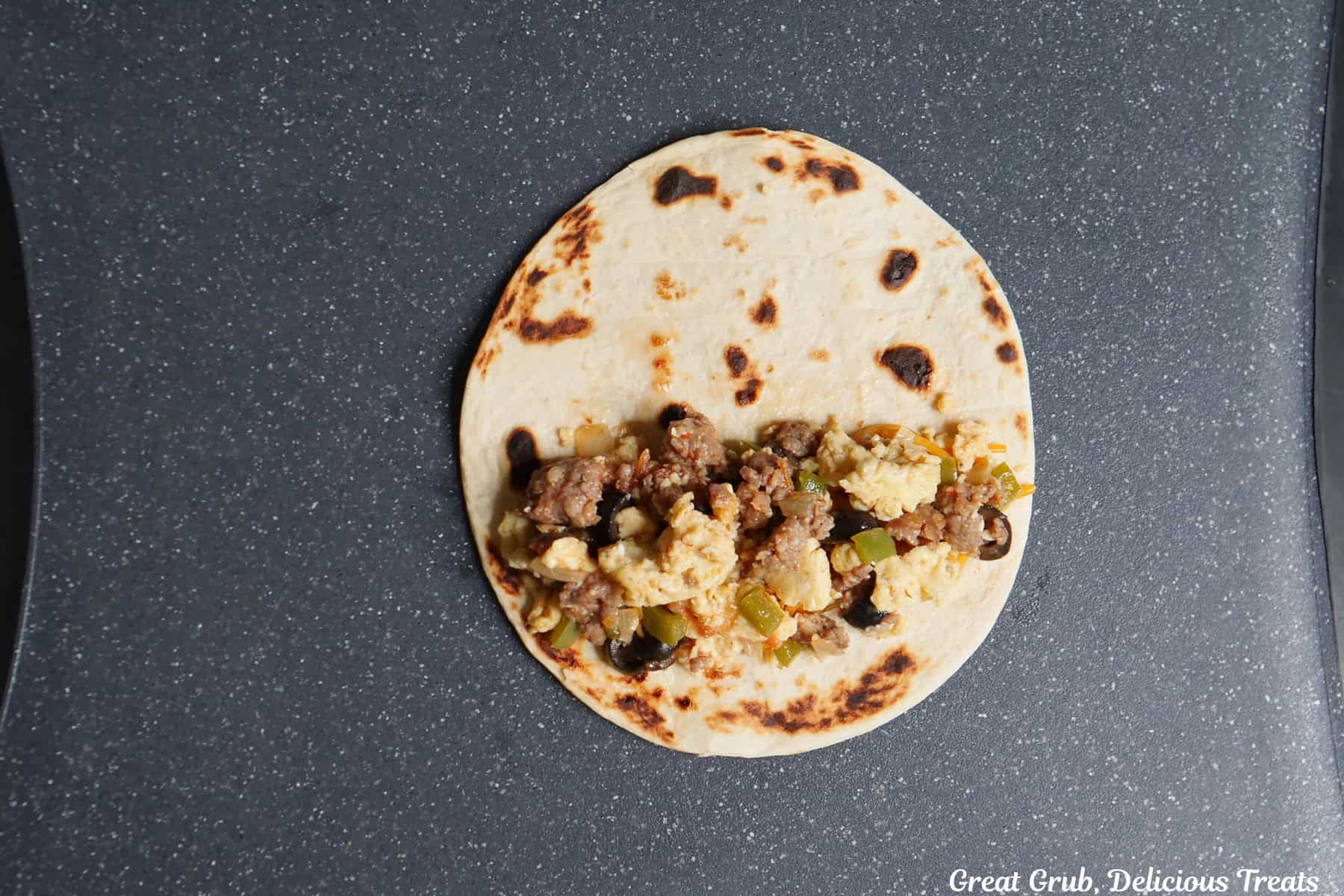 A black surface with a flour tortillas on it with shredded cheese, sausage and egg mixture, and is about to be rolled up.