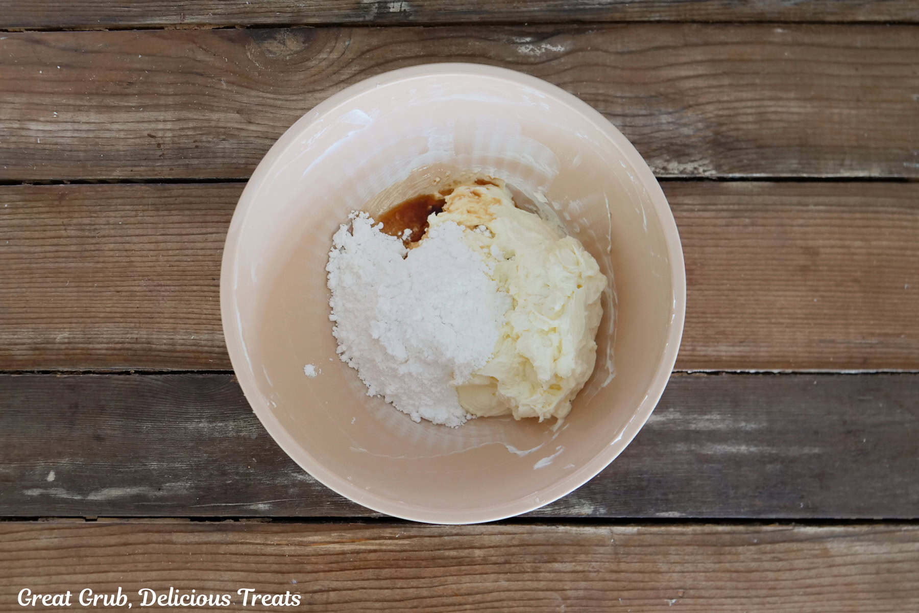 A rustic wood surface with a cream color bowl filled with cream cheese, powdered sugar, and vanilla in it.