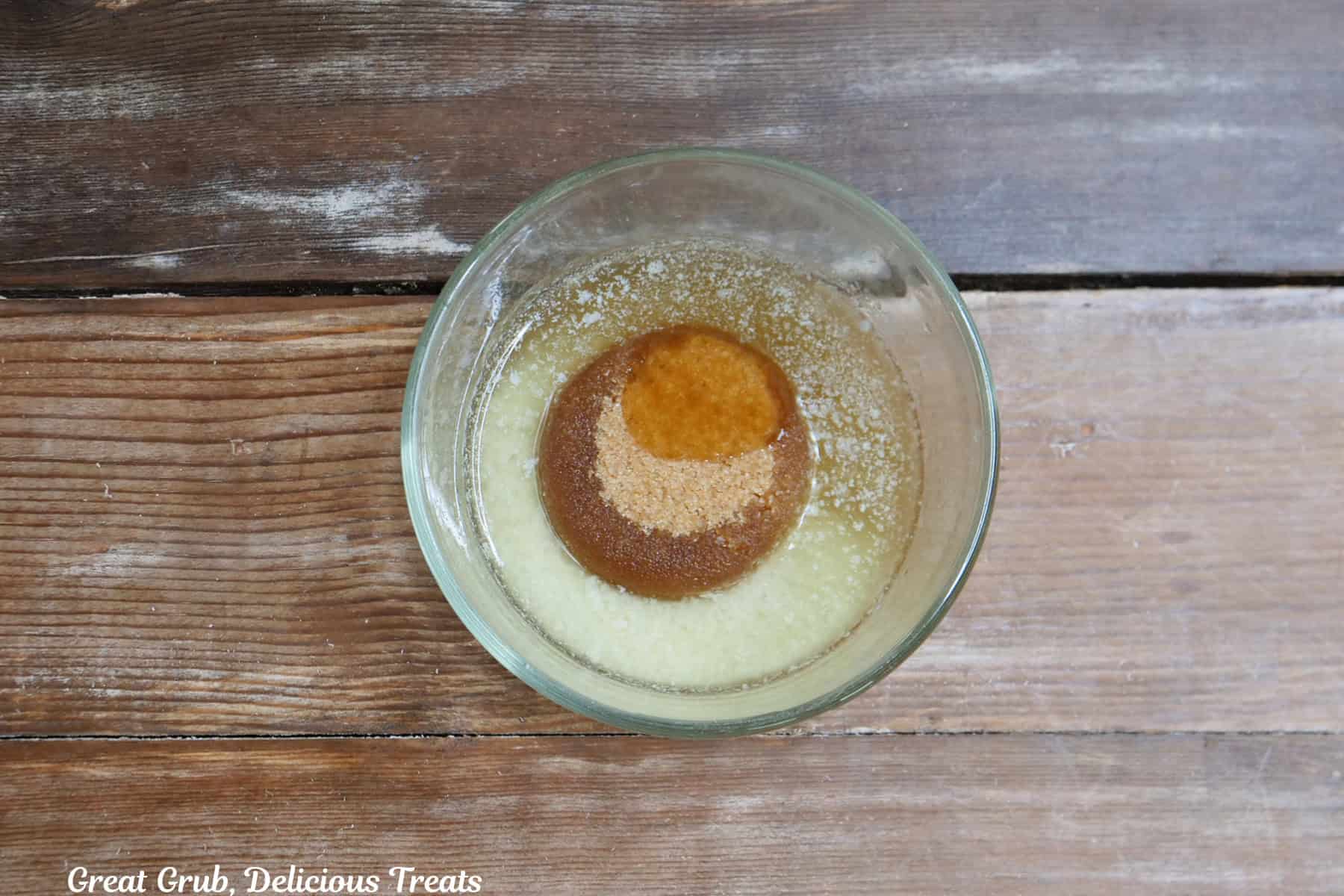 A rustic wood surface with a glass bowl filled with melted butter, brown sugar, and honey before being mixed together.