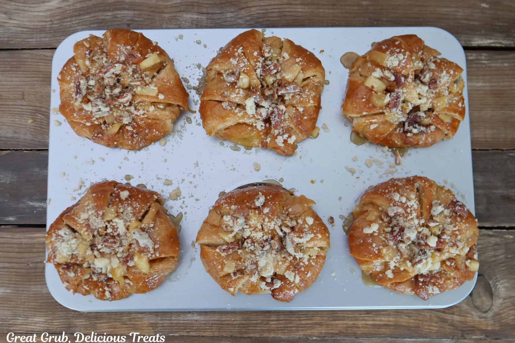 A wood surface with a silver baking tray with six apple cheesecake croissants on it and about to be put in the oven to bake.