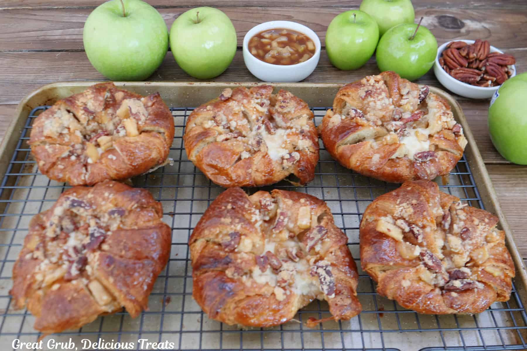 A baking sheet with a wire rack on top with six apple cheesecake croissants on it with green apples, apple filling, and pecans in the background.