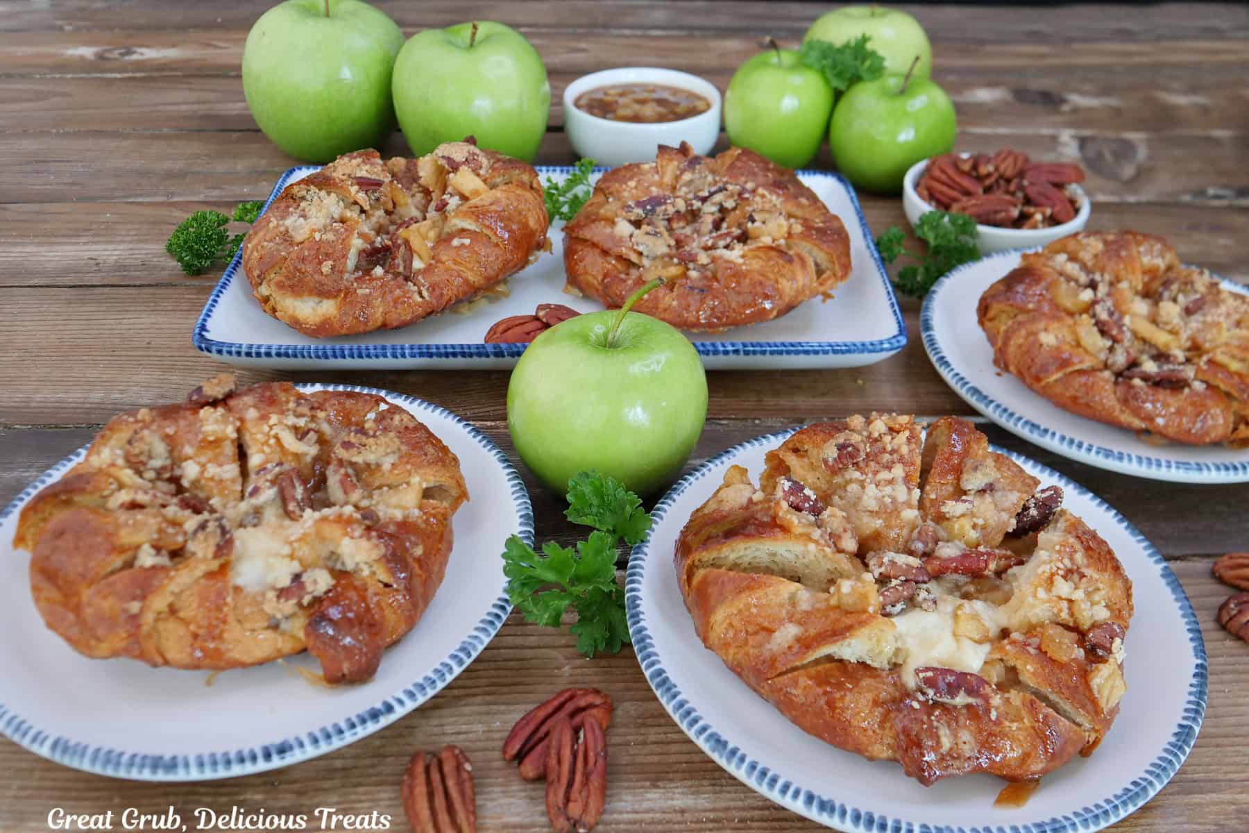 A rustic wood surface with four white plates with blue trim with apple cheesecake croissants on them with green apples in the background.