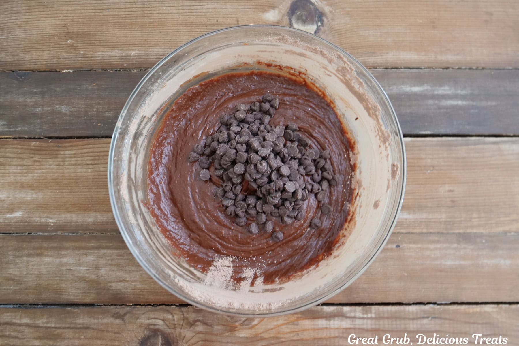 A wood surface with a glass bowl with the brownies batter in it and chocolate chips added to the bowl.