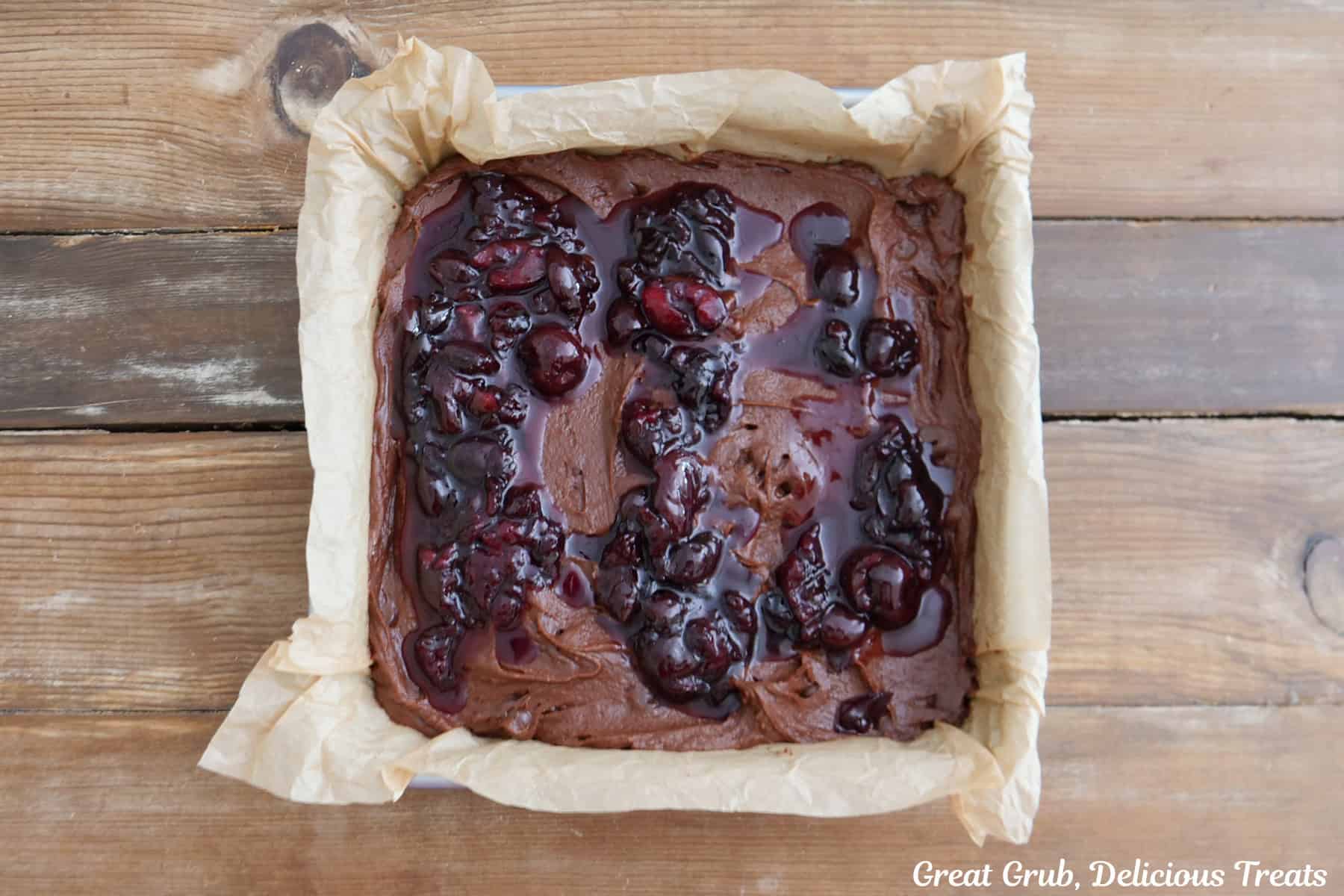 A wood surface with an 8 by 8 baking pan lined with parchment paper with the black forest brownie batter in it and topped with the cherry topping.