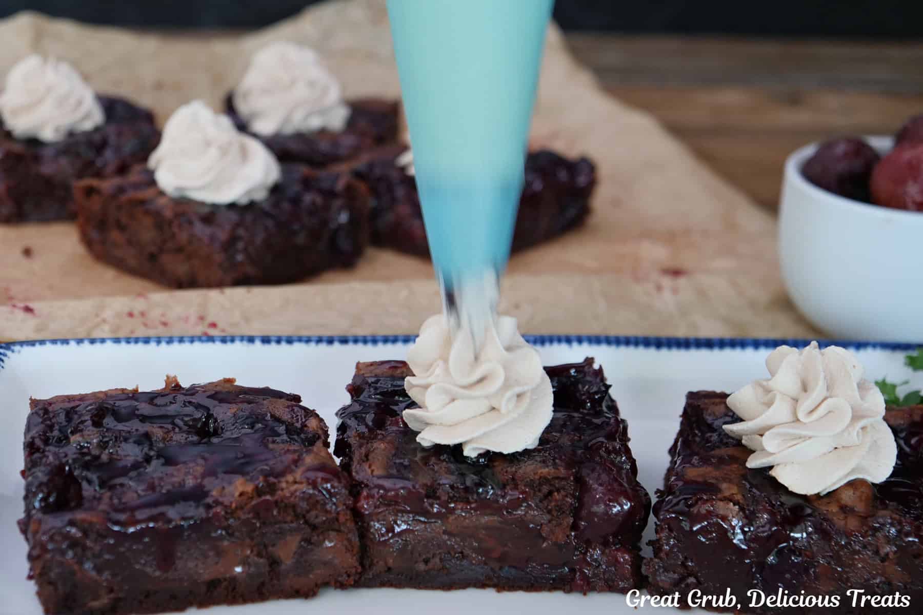 A white oblong plate with blue trim with three brownies on it being topped with whipped cream.