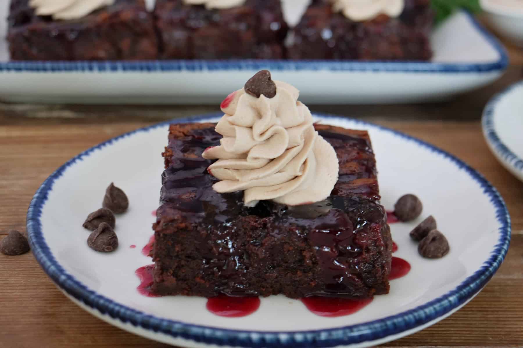 A wood surface with a white plate with blue trim and a brownie on it topped with whipped cream and chocolate chips placed around the plate.