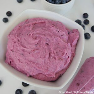 A white surface with a white bowl filled with homemade blueberry buttercream frosting in it with a piping bag filled with frosting, and fresh blueberries scattered around the white surface.