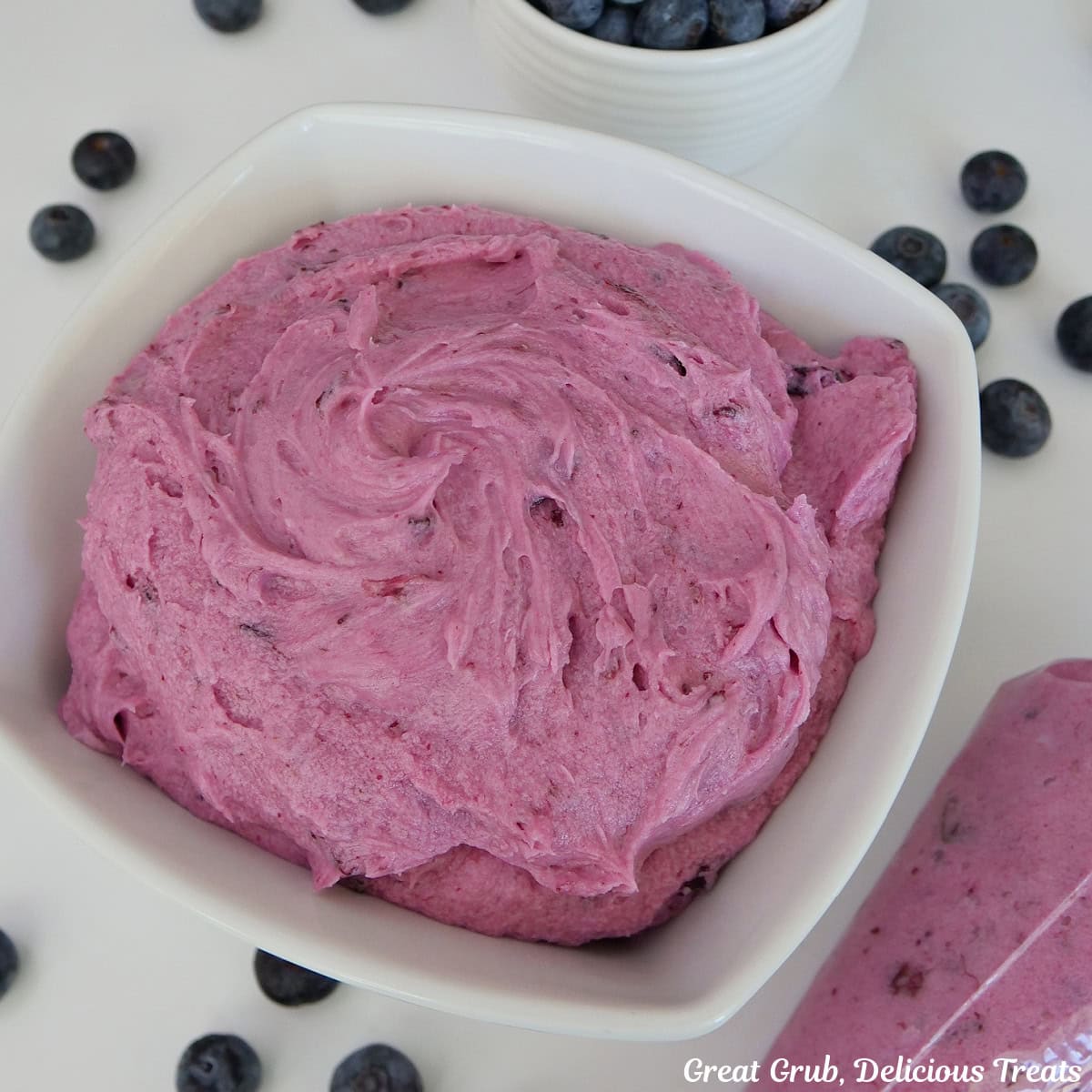 A white surface with a white bowl filled with homemade blueberry buttercream frosting in it with a piping bag filled with frosting, and fresh blueberries scattered around the white surface.
