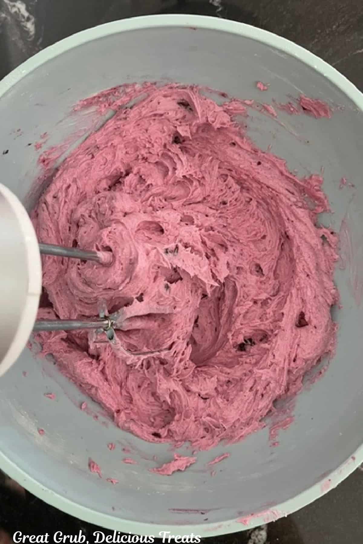 A light blue bowl with blueberry buttercream frosting in it after being mixed together with the hand mixer beater attachments still attached.