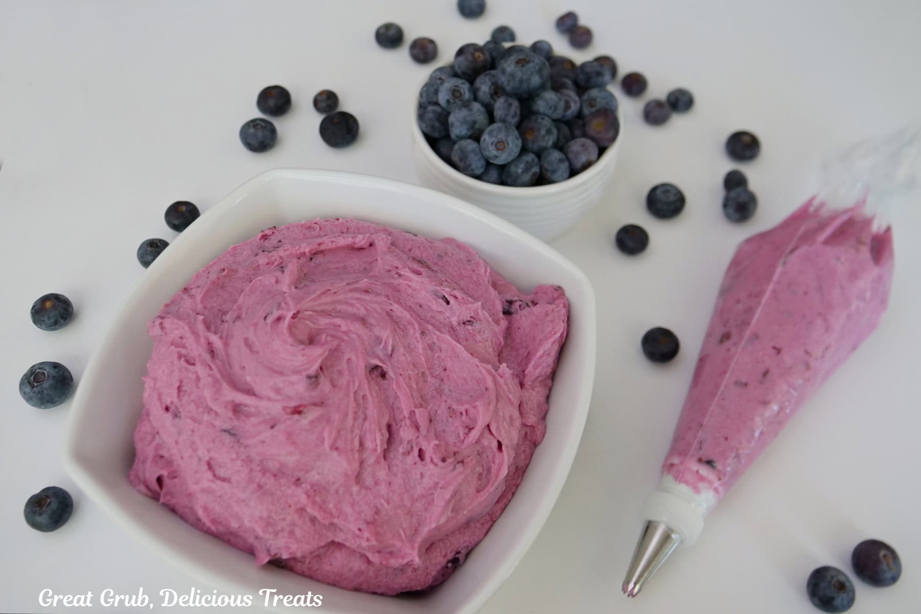 A white surface with homemade blueberry buttercream frosting in a white bowl and in a clear piping bag, with a small white bowl filled with fresh blueberries, and some blueberries scattered around the surface.