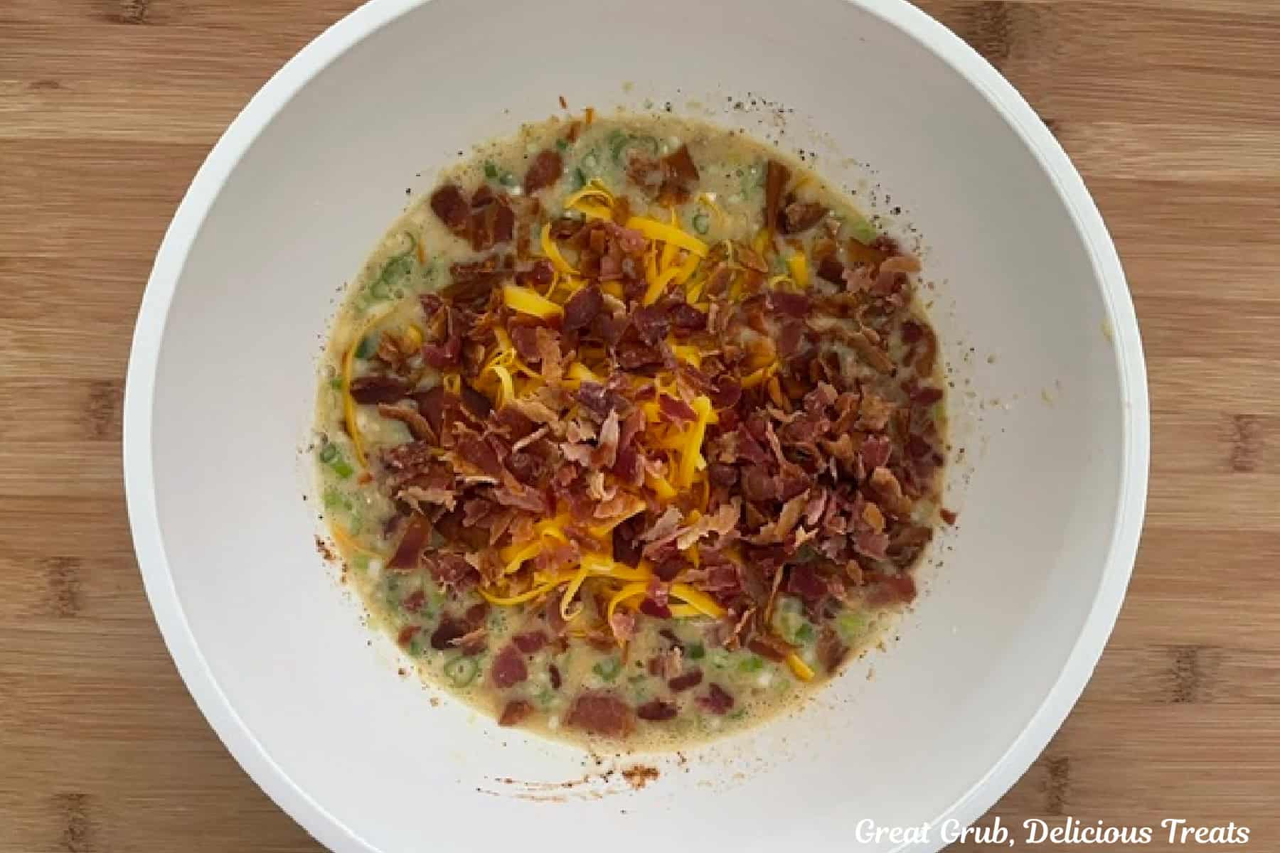 A wood surface with a white bowl filled with all the ingredients in it to make cheesy egg bites.