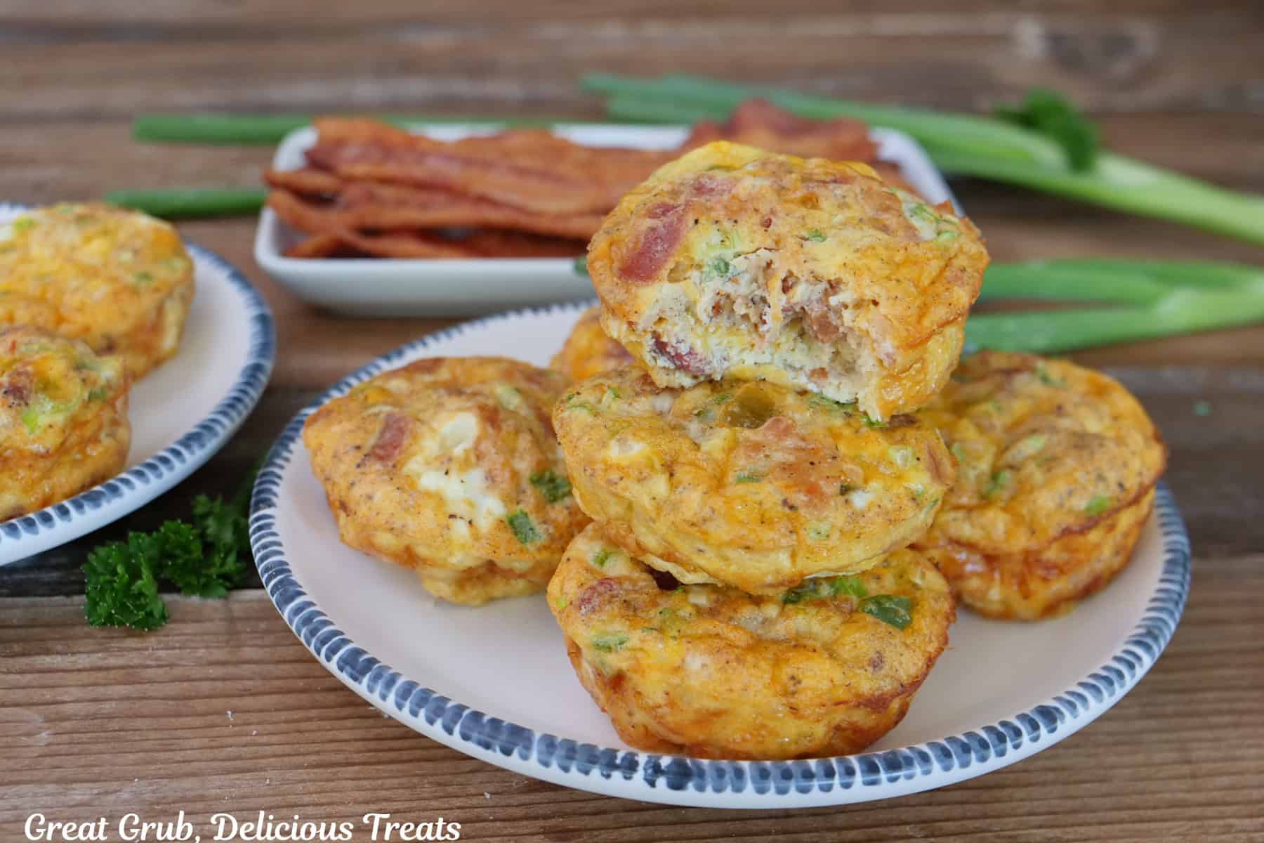 A wood surface with two white plates with blue trim with cheesy egg bites stacked on top of each other with a bite taken out of the one on top, with bacon and green onions in the background.
