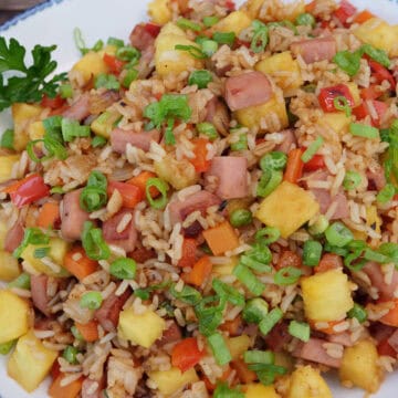 A wood surface with a white bowl with blue trim filled with Hawaiian fried rice with pineapple and Spam.
