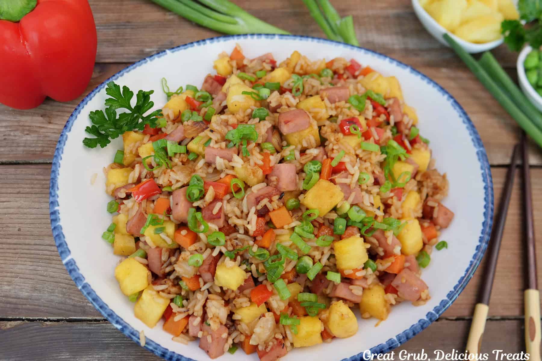 A wood surface with a blue trimmed white bowl filled with Hawaiian fried rice with pineapple and Spam.