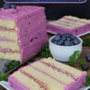 A wood surface with a white plate with a slice of a four layer lemon cake with blueberry frosting on it with the cake and another slice in the background.