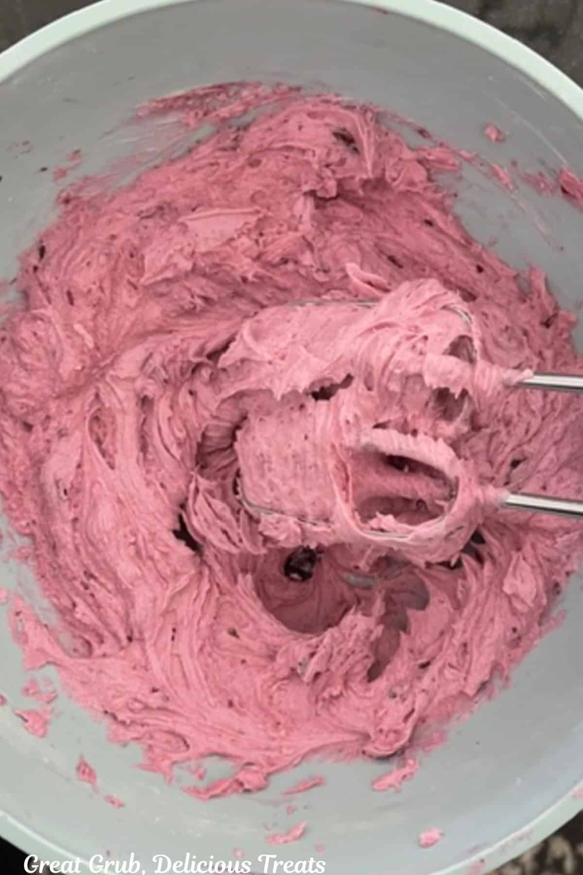 A light blue bowl with blueberry buttercream frosting being beat with the mixer beaters in the bowl.