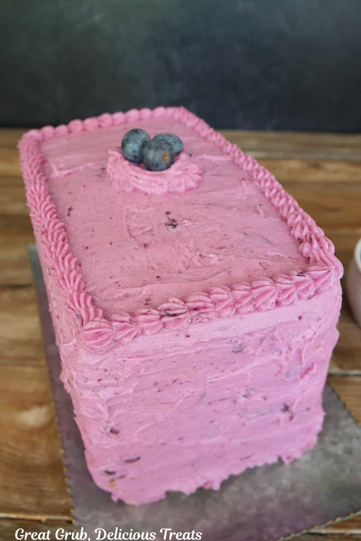 A wood surface with a sliver rectangle tray with a blueberry buttercream frosted four layer cake on it.