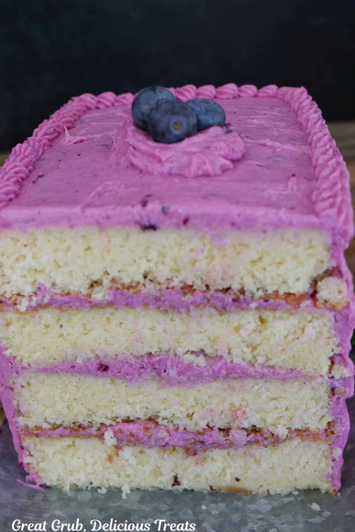 A four layered lemon cake with blueberry buttercream frosting between each layer and on the top and sides of the cake.