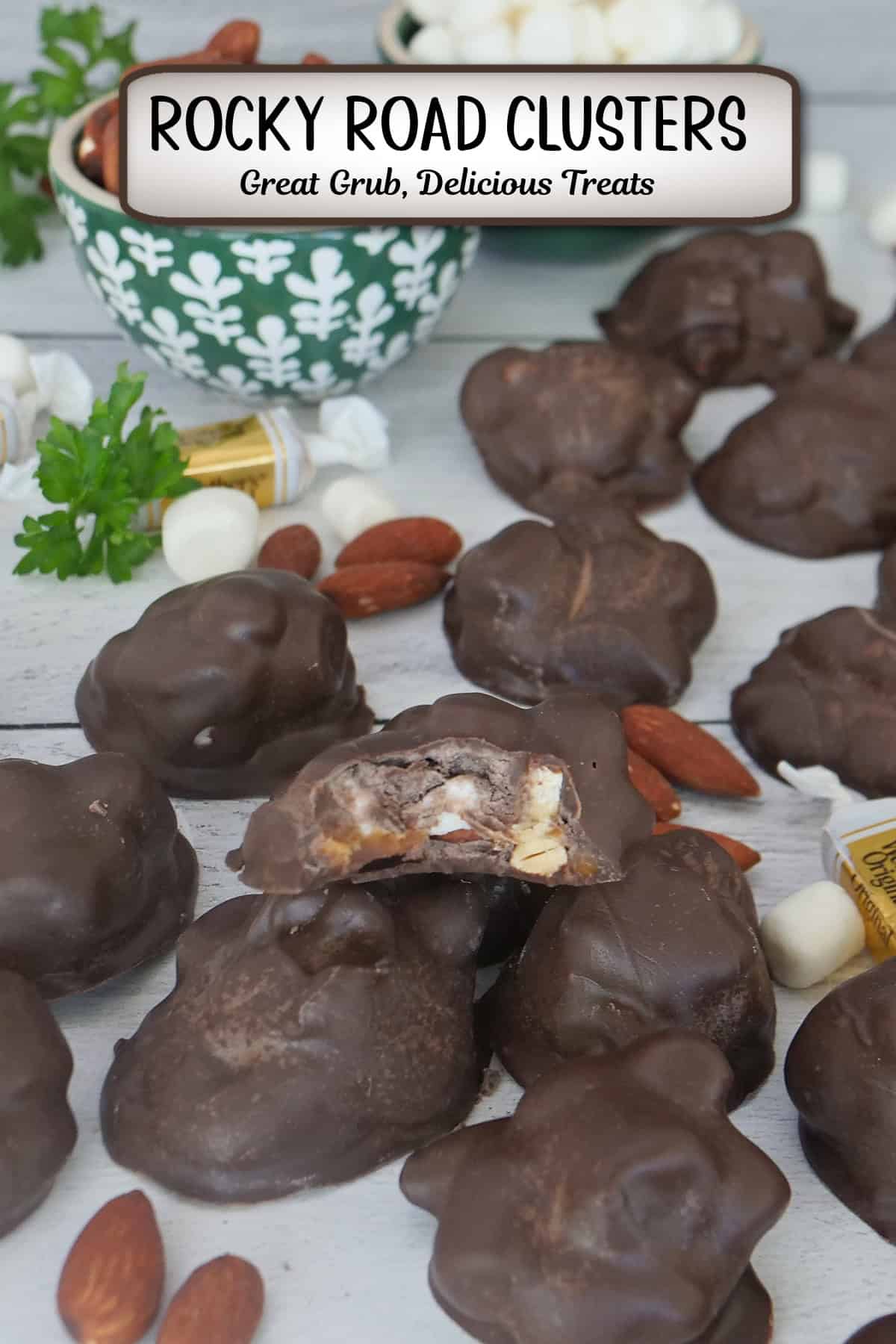A white surface with homemade chocolate covered rocky road clusters placed randomly all over and a bite taken out of one of the clusters, and a title overlay at the top.