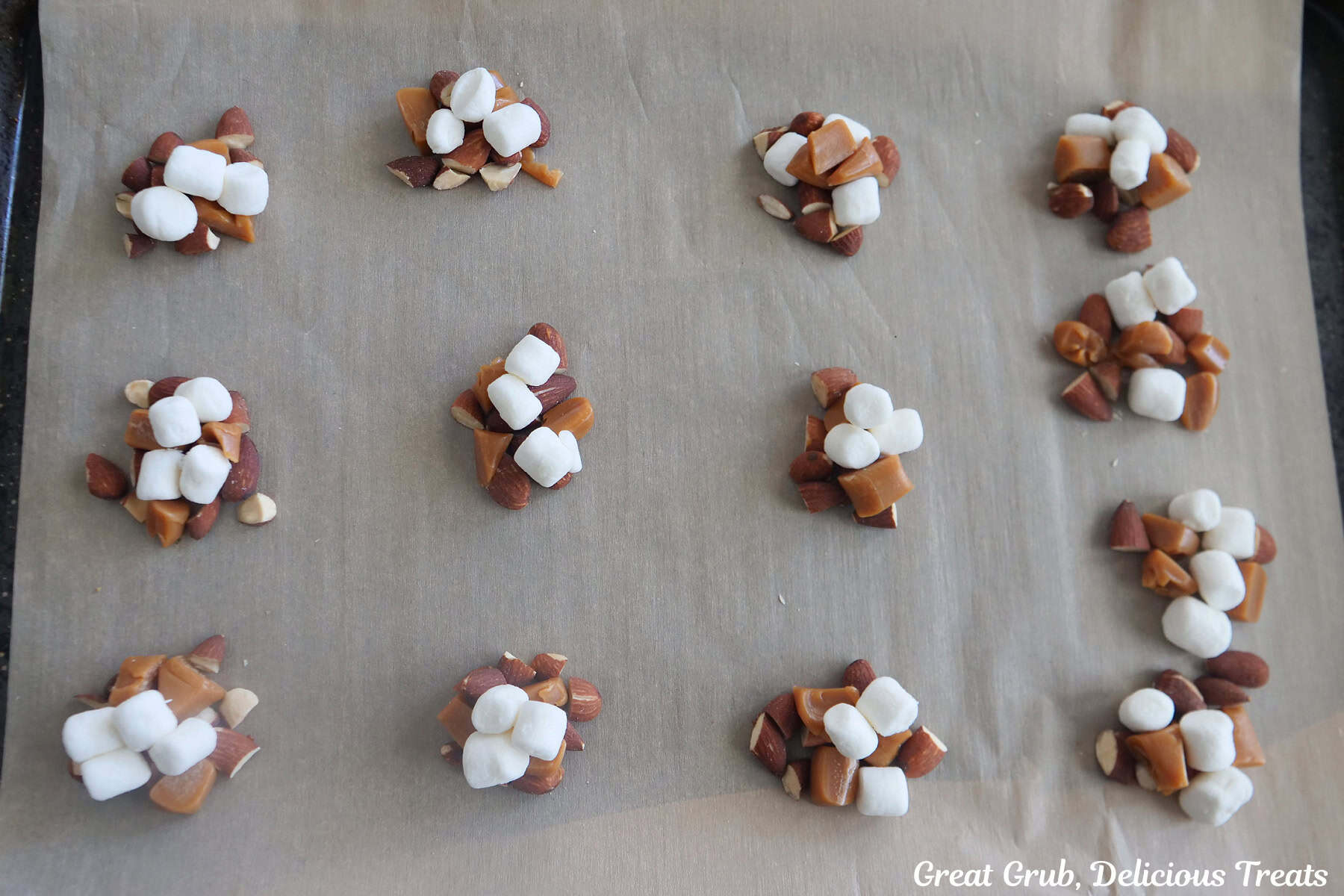 A parchment lined baking sheet with twelve clusters of caramel, mini marshmallows, and almonds placed on it.