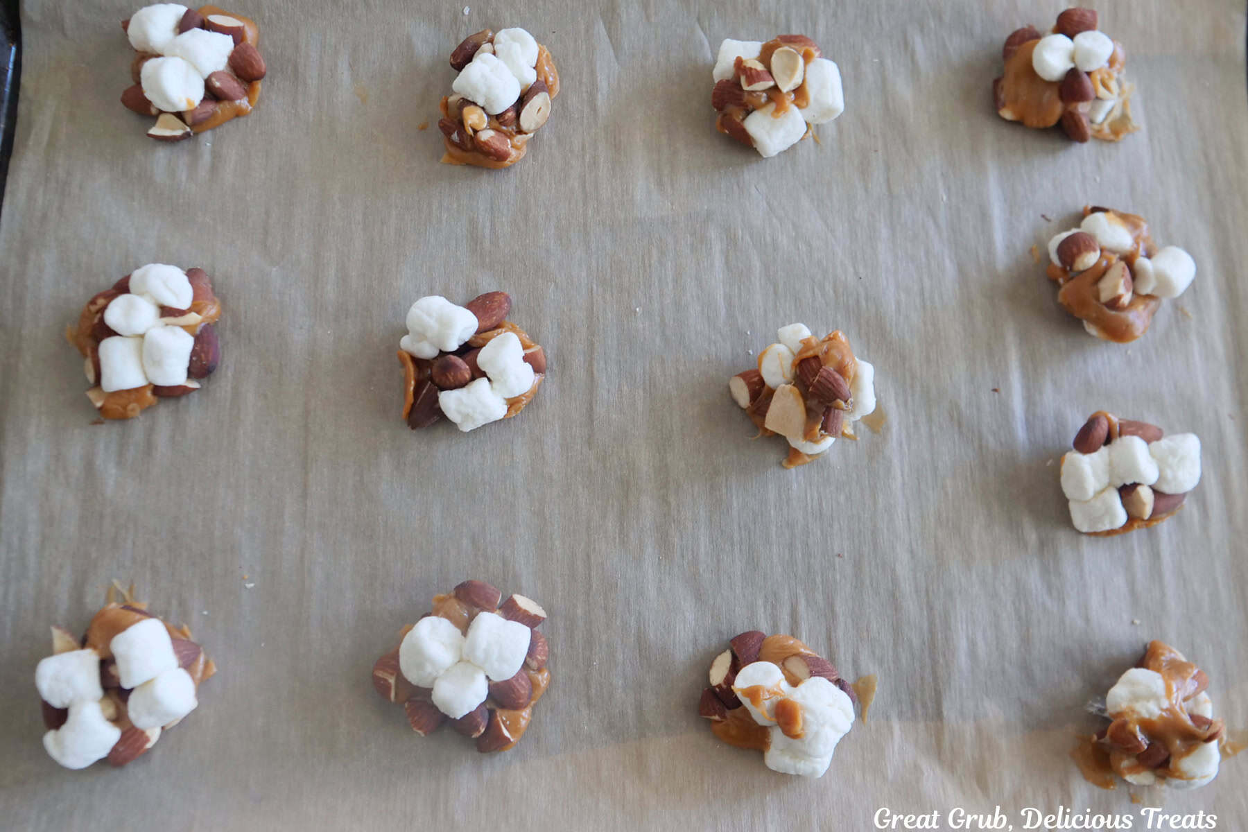 A parchment lined baking sheet with twelve clusters of mini marshmallows, almonds, and caramel after it was heated up in the oven just until the caramel started to melt and held everything together.