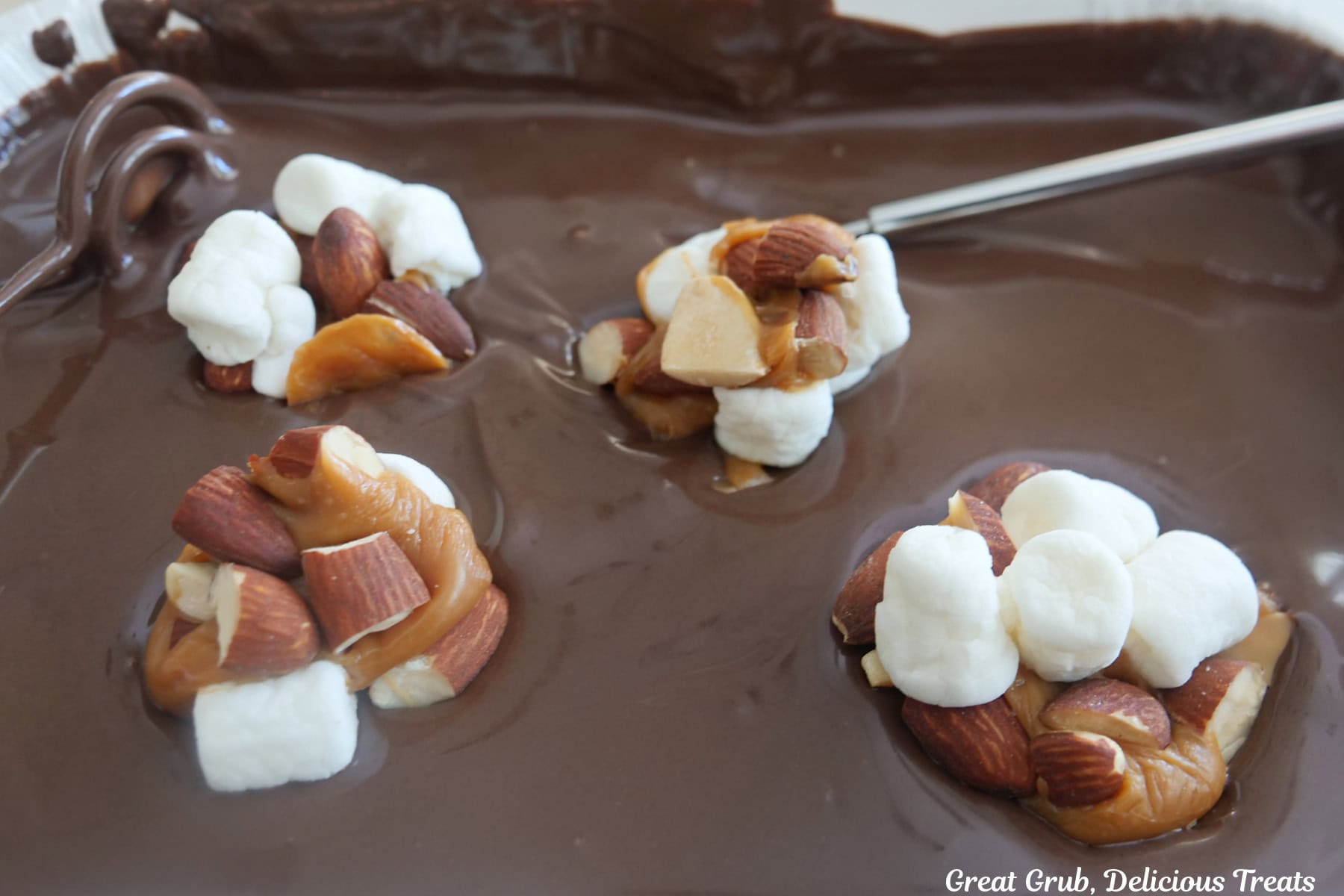 A white tray with melted chocolate in it with four rocky road clusters placed in it to be covered in the melted chocolate.