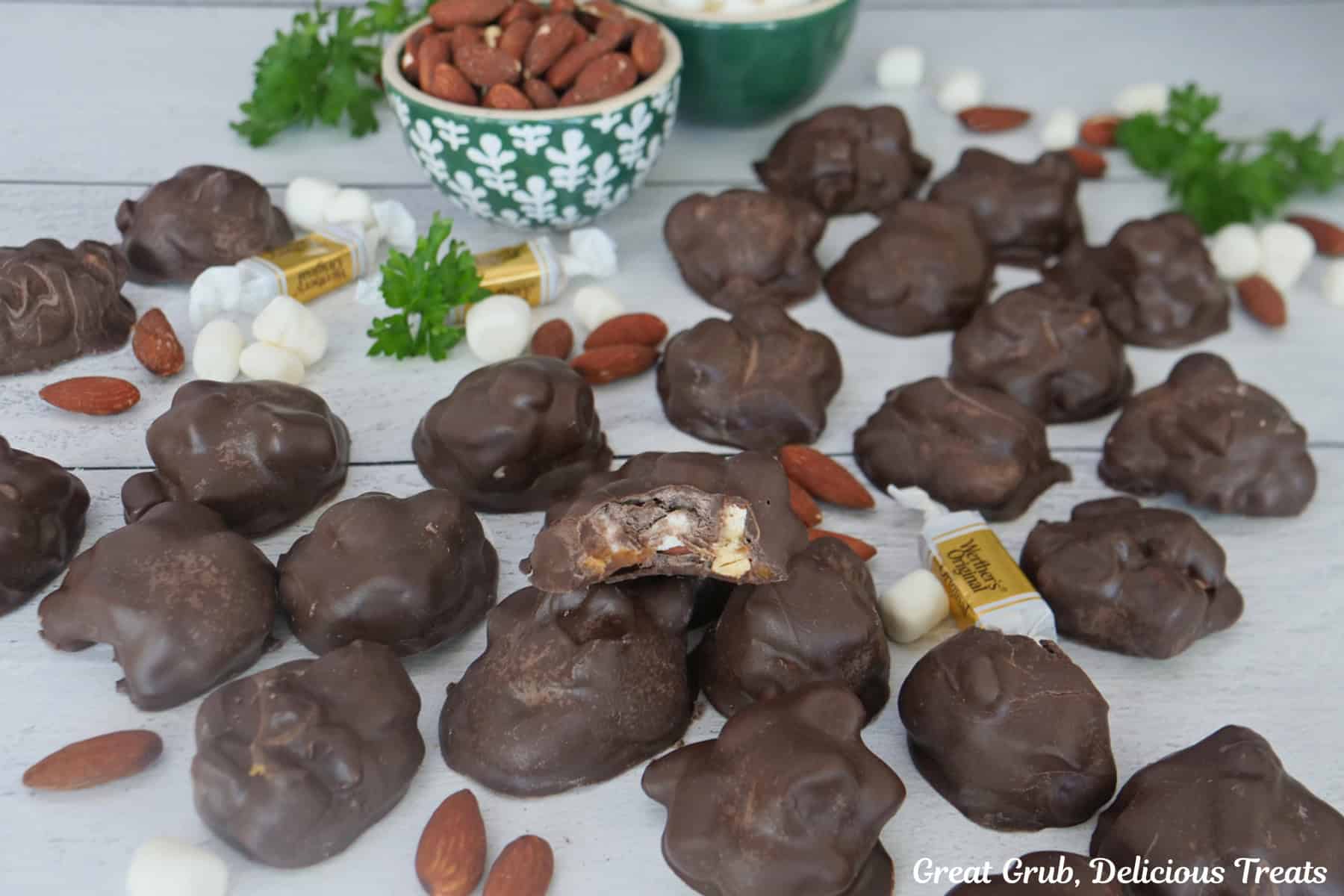 A white surface with two dozen pieces of rocky road candy clusters placed on it along with caramel, almonds, and mini marshmallows.
