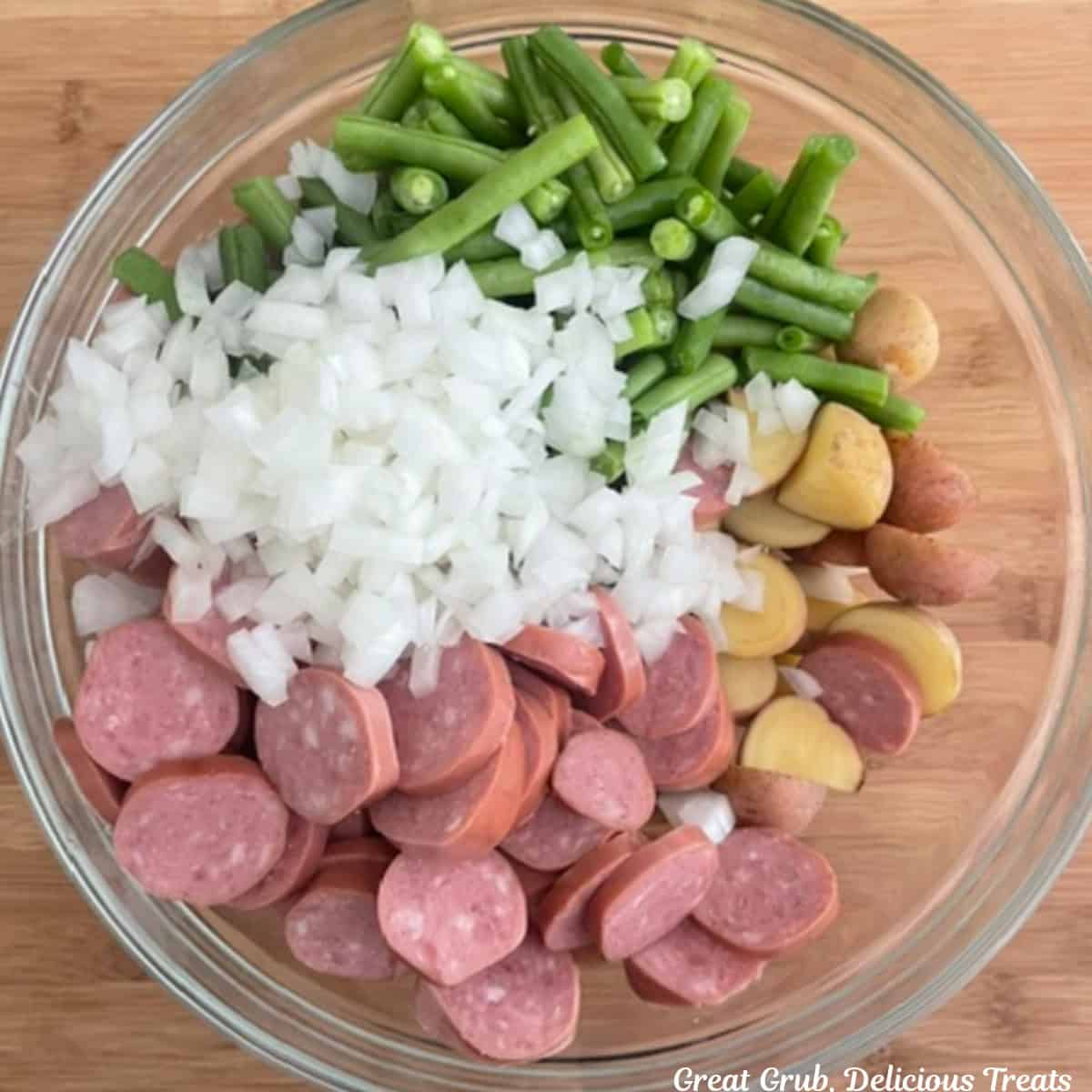 A wood surface with a glass bowl filled with sliced sausage, baby creamer potatoes, fresh green beans, diced onions, and garlic.