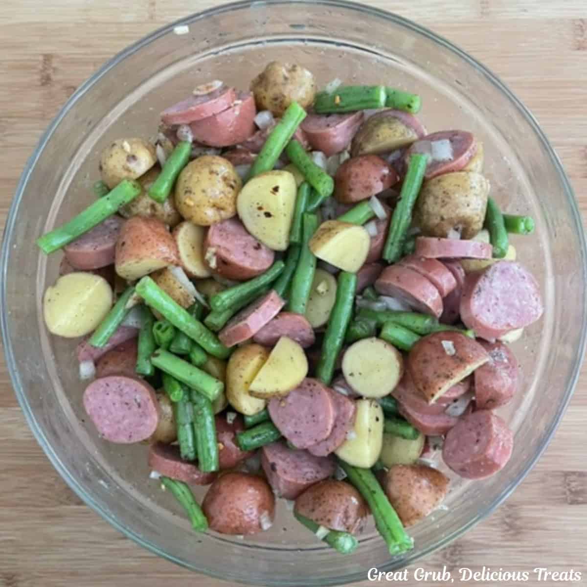 A glass bowl placed on a wood surface filled with sliced sausage, potatoes, green beans, onions, garlic, and seasonings all mixed together.