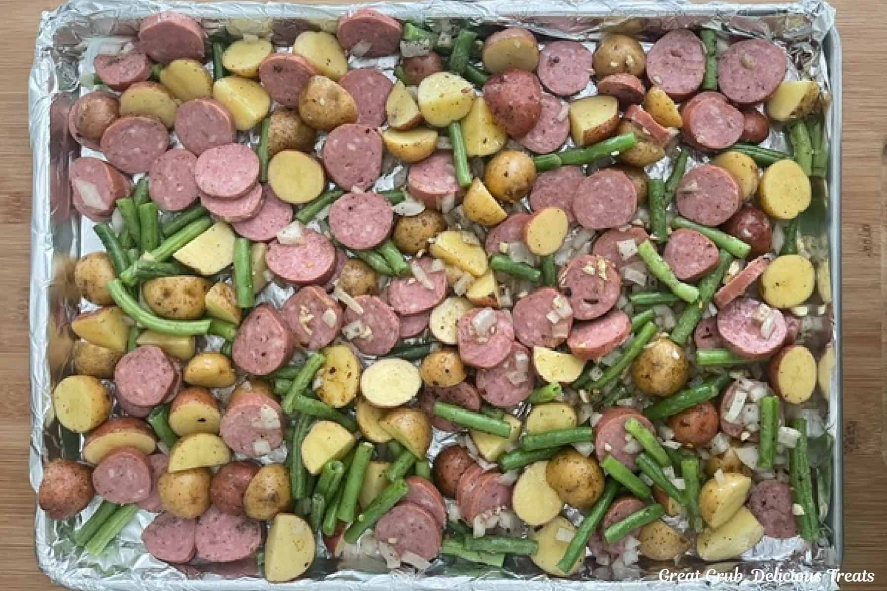 A foil lined large baking sheet with sliced sausage, green beans, cut potatoes, onions, garlic, seasonings placed on it before being baked.