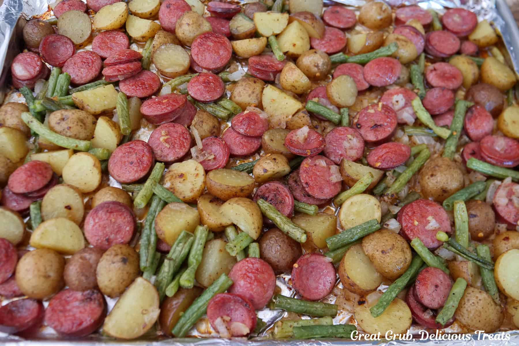 A large foil lined baking sheet with cooked sausage, green beans, potatoes, onions, and seasonings.