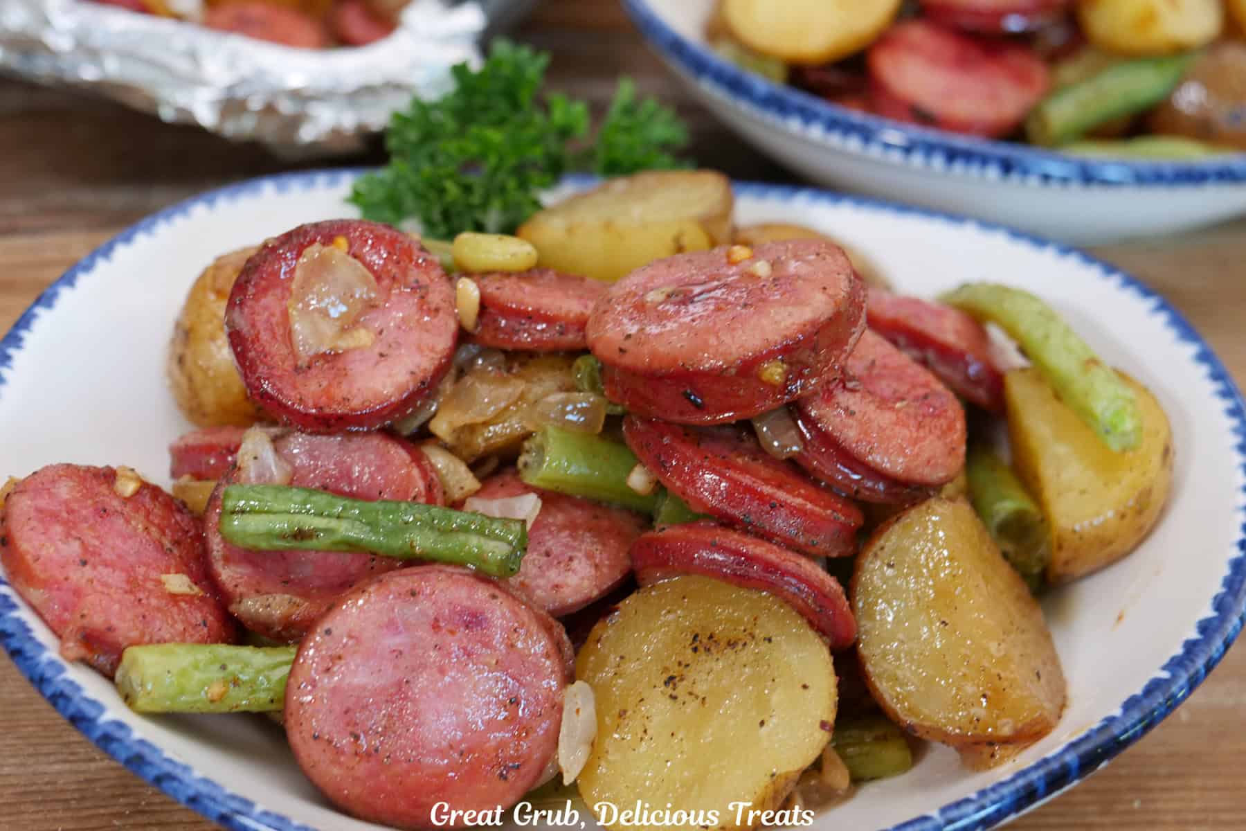 A wood surface with a white bowl with blue trim filled with a serving of smoked sausage, potatoes and green beans with another filled bowl in the background.