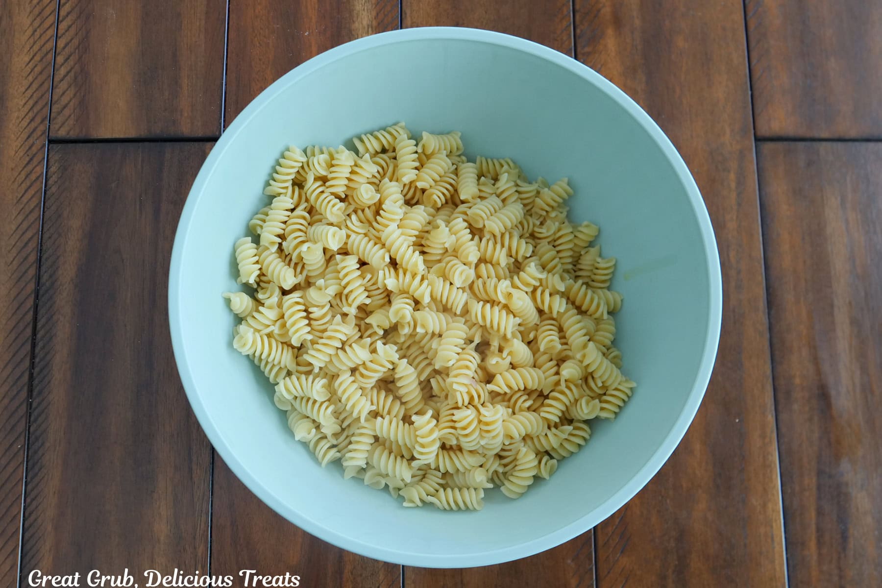 A wood surface with a light blue bowl with cooked rotini pasta in it.